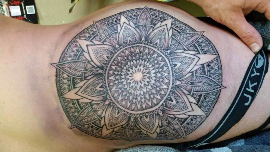 A person displays a large, detailed mandala tattoo on their back, characterized by intricate geometric and floral patterns.