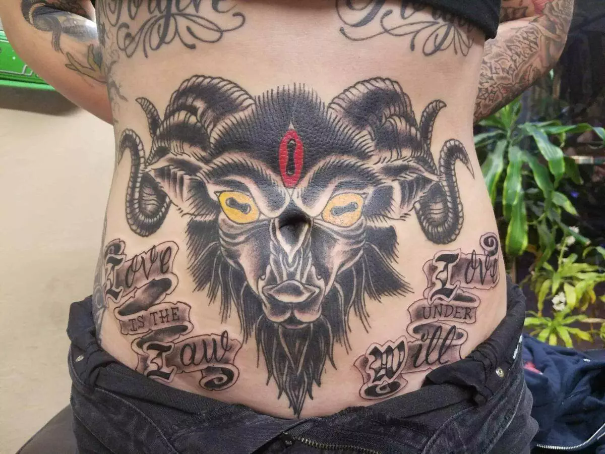 A person with a detailed tattoo of a goat with prominent horns and yellow eyes on their torso, featuring text that reads 