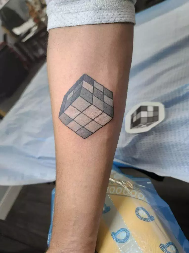 A person's forearm with a tattoo of an incomplete Rubik's Cube showing varying shades of black and gray. A printed design of the same tattoo is seen nearby on a blue sheet.