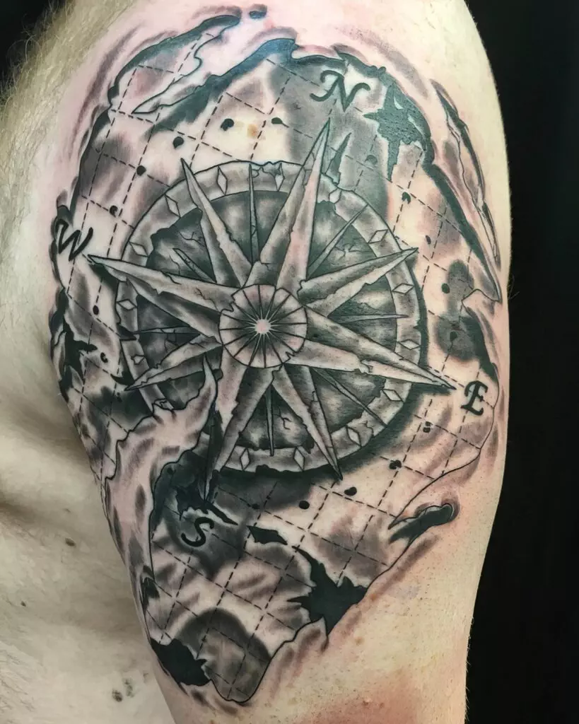 A tattoo featuring a detailed compass rose surrounded by a shaded, abstract background, located on an individual's upper arm.