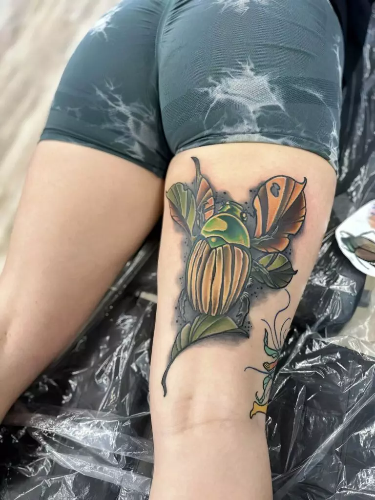 Tattoo of a colorful beetle and leaves on the back of a person's thigh, while they are lying face down wearing dark shorts.