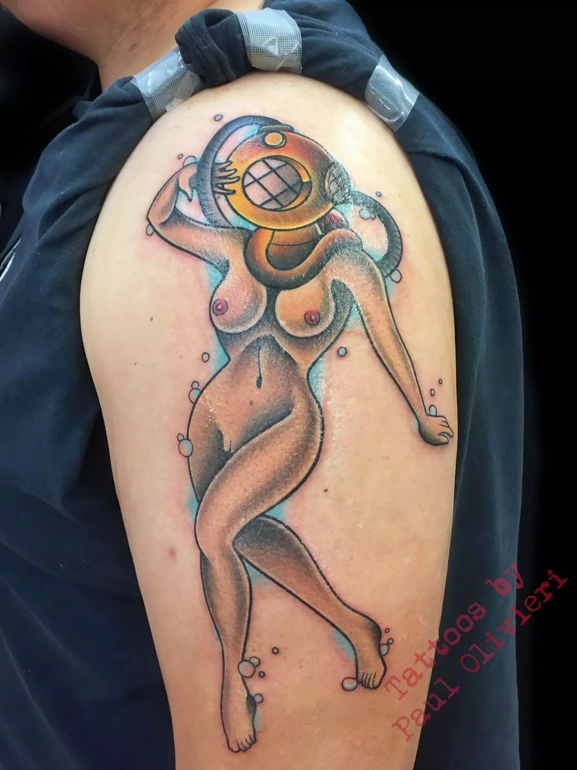 A tattoo of a stylized nude woman with a vintage diving helmet on her head, located on a person's upper arm. The image features the text 