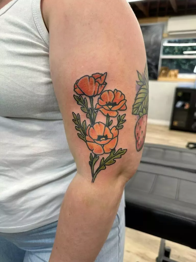 A person's arm with a tattoo of orange flowers and green leaves. They are wearing a white tank top and light blue pants, and the background shows a tattoo studio setup.