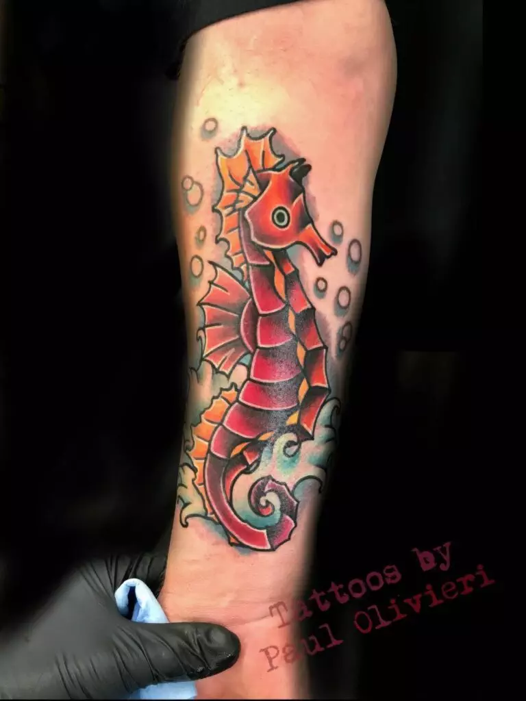 A colorful seahorse tattoo on a person's forearm with bubbles surrounding it. A gloved hand is seen holding a cloth at the bottom of the arm. The text 