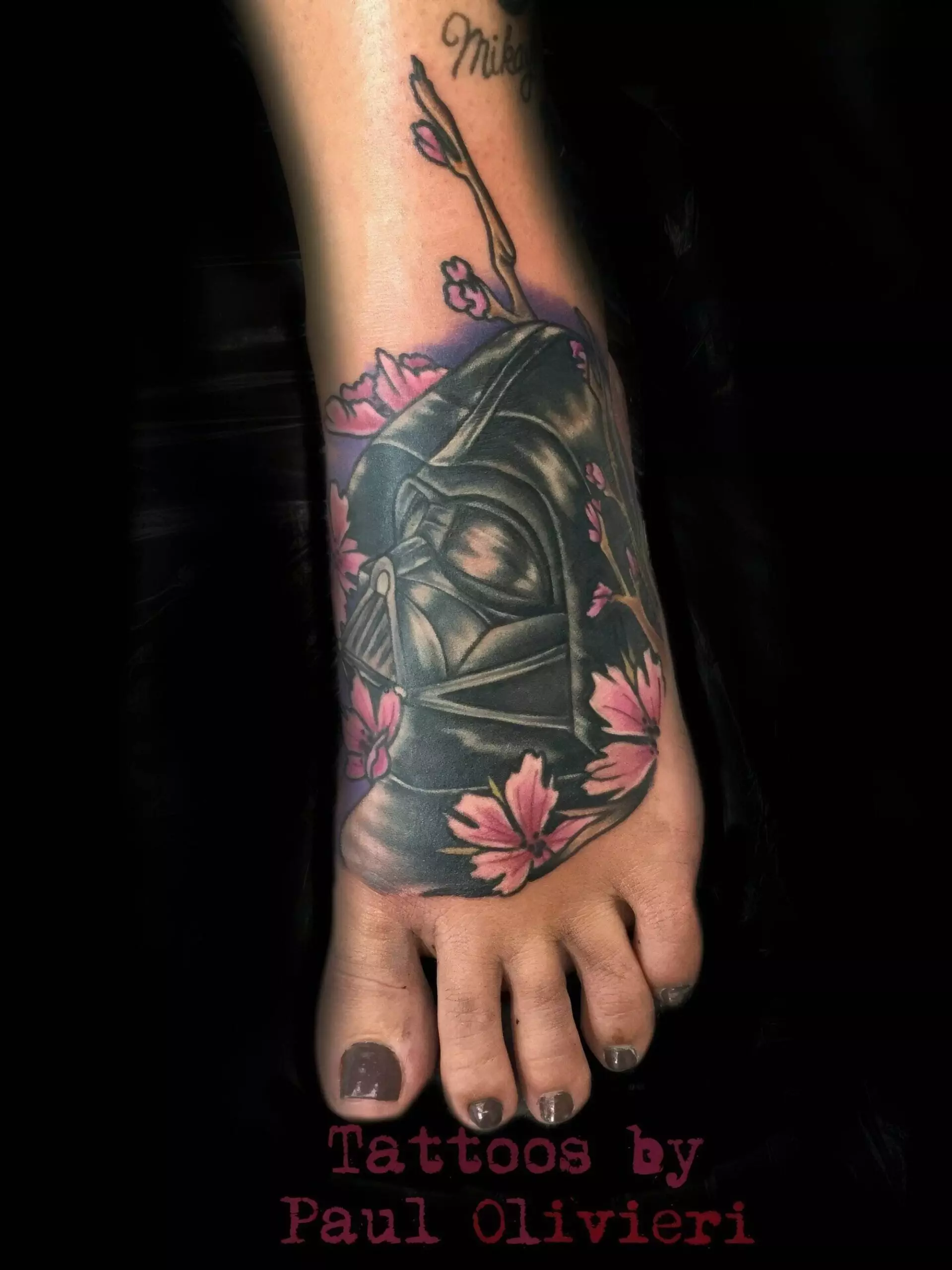 A foot tattoo depicting a helmeted figure surrounded by pink flowers, with text at the bottom reading 