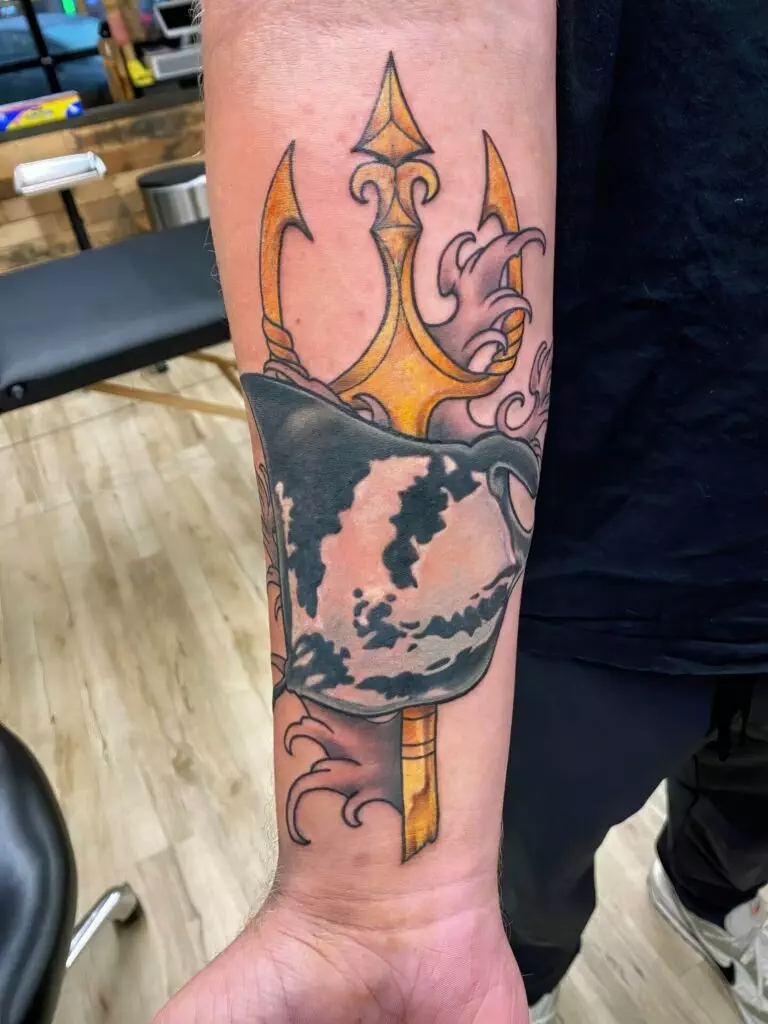 Forearm tattoo depicting a black helmet with golden details and a trident overlaid with swirling patterns. The background features a blurred view of what appears to be a tattoo studio.