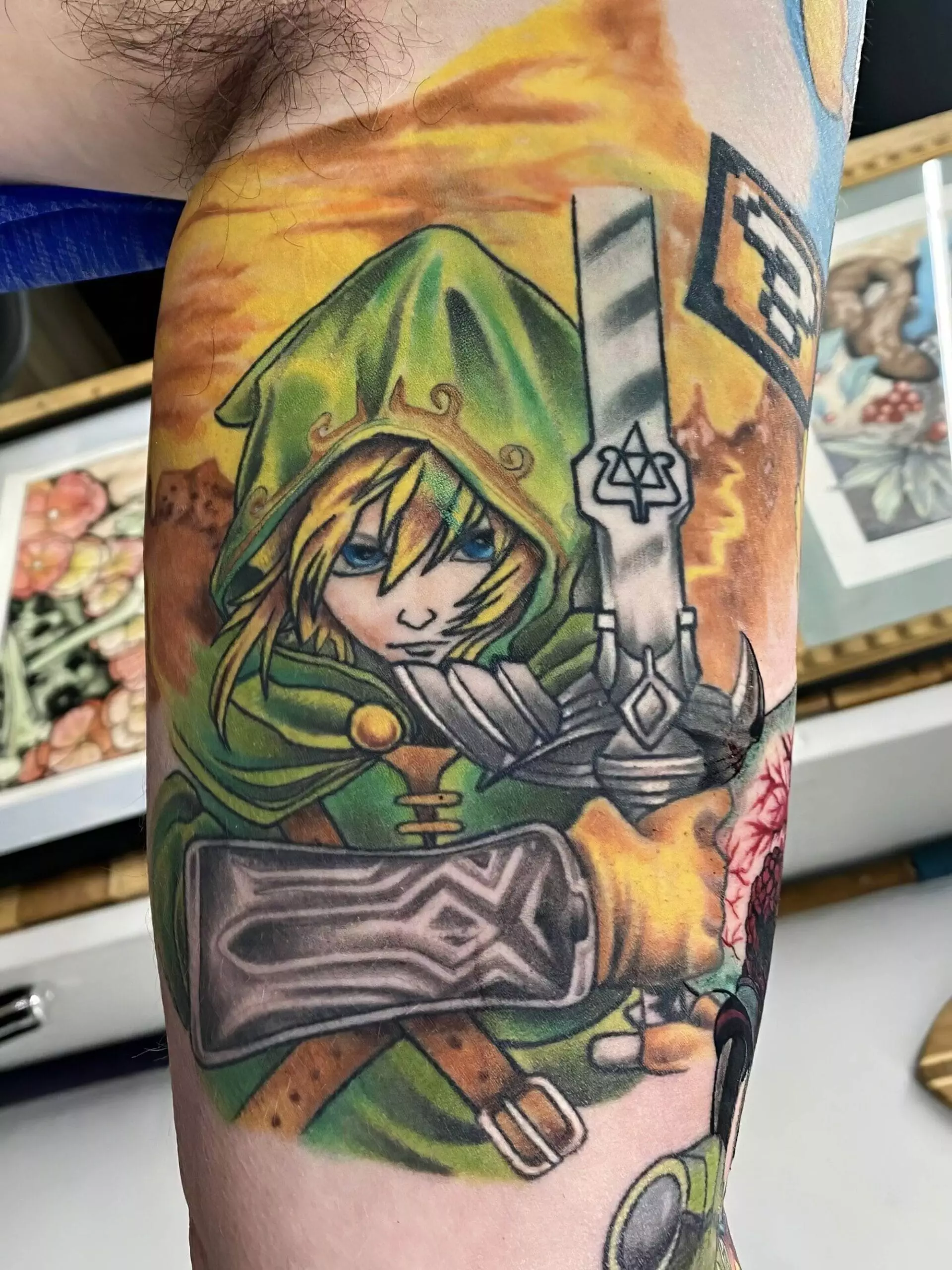 A detailed tattoo featuring a character in a green hood with a sword, surrounded by vibrant colors and artistic background elements.