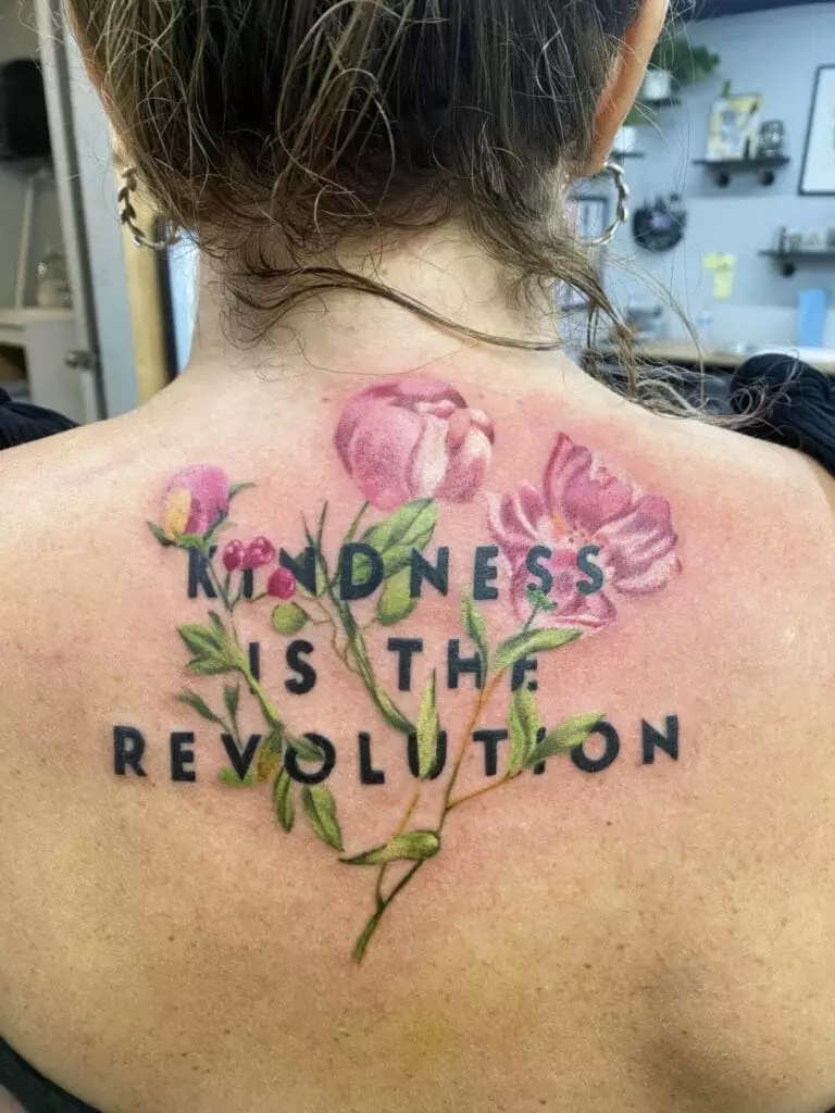 A tattoo on a woman's upper back features pink flowers intertwined with the words 