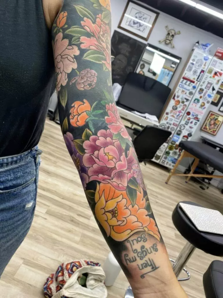 A person with a colorful floral tattoo sleeve on their arm, featuring various flowers and leaves, stands in a tattoo studio. The text 