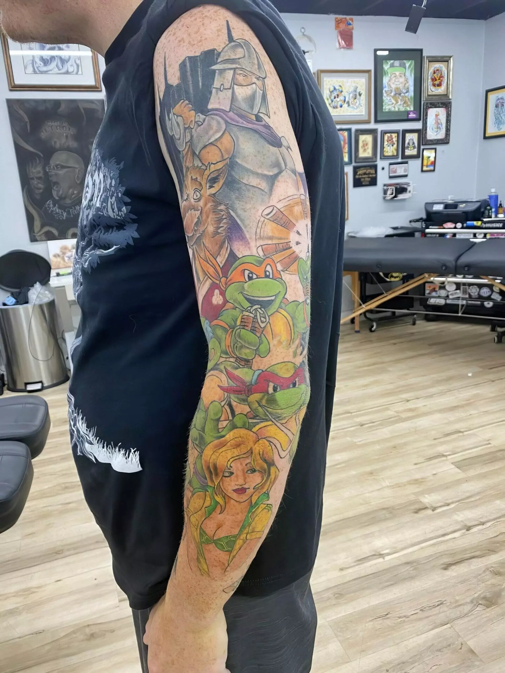 A person displays a colorful arm tattoo featuring various characters, including warriors and cartoon figures, in a room with framed artwork on the walls.