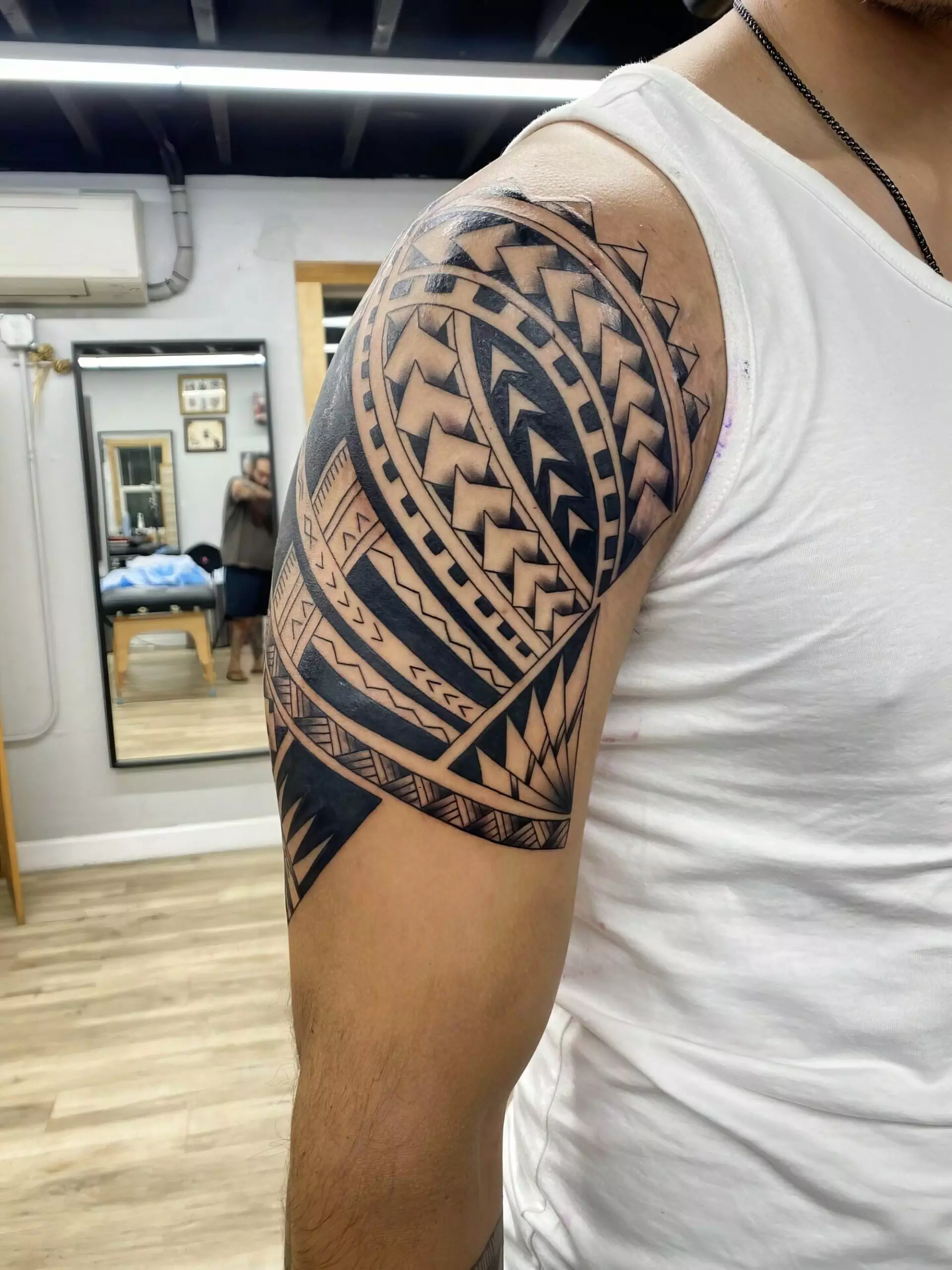 A close-up of a person's upper arm showcasing a detailed black geometric tattoo, including patterns of triangles, lines, and curves. The person is wearing a white sleeveless shirt.