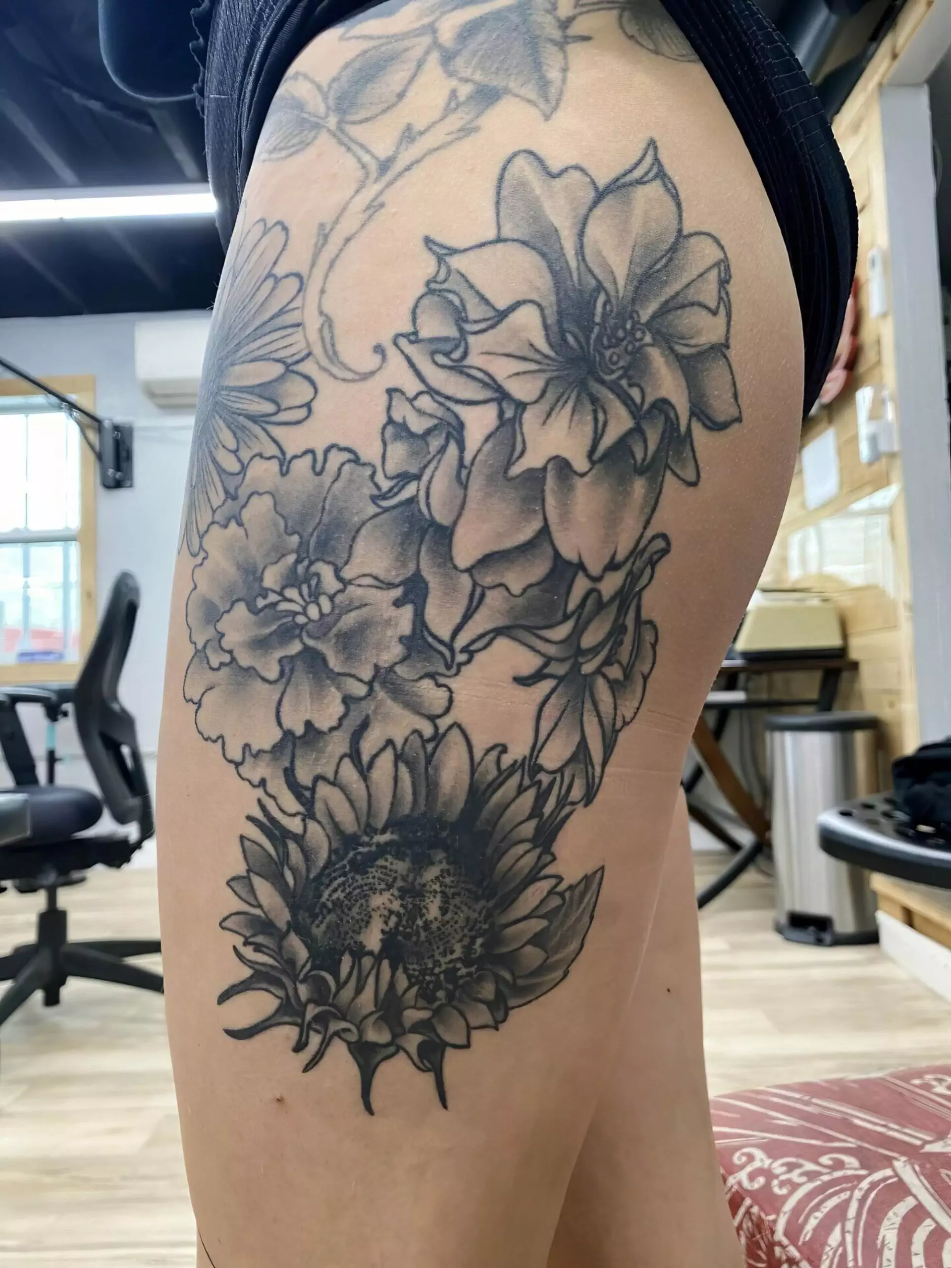 A thigh tattoo showcasing a detailed black and grey floral design, including various flowers, is visible. The person is wearing black shorts, and the background features an indoor setting with furniture.