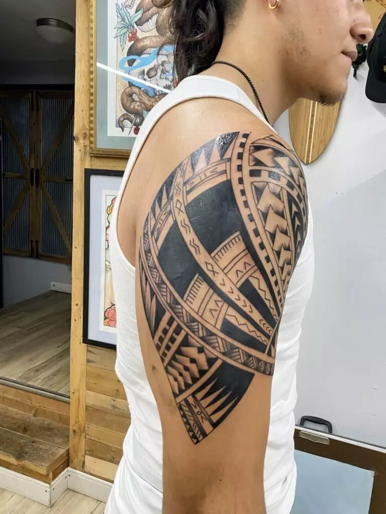 A person wearing a white tank top shows a detailed large black geometric tattoo on their upper arm and shoulder. Various framed artworks are visible in the background.