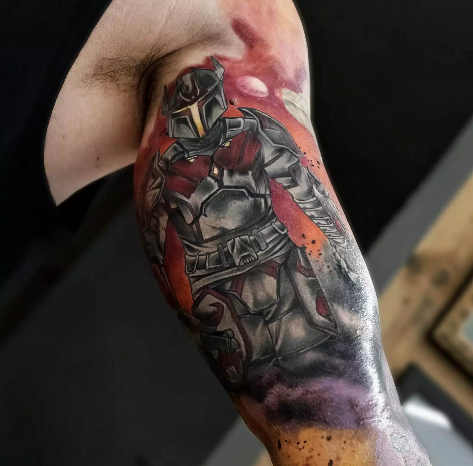 Detailed Mandalorian character tattoo on a person's upper arm in black and red ink with a colorful background.