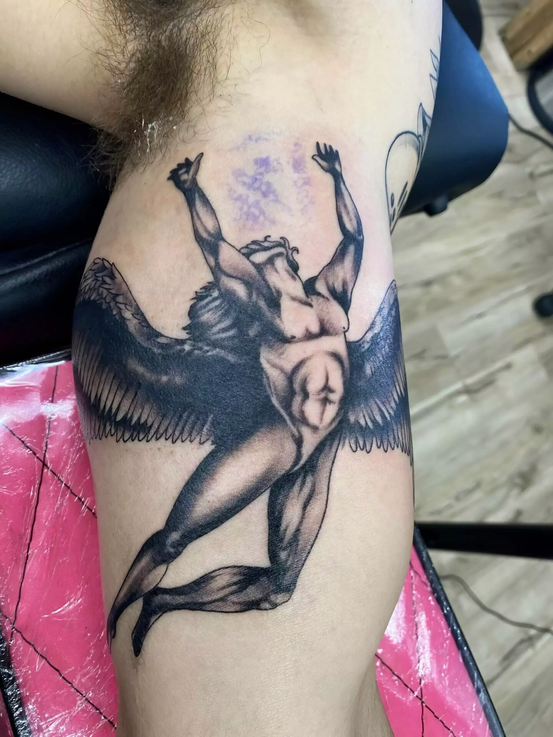 A detailed black and gray tattoo on an arm depicting a muscular winged figure reaching upward, with detailed shading and muscle definition. The arm rests on a pink and black surface.