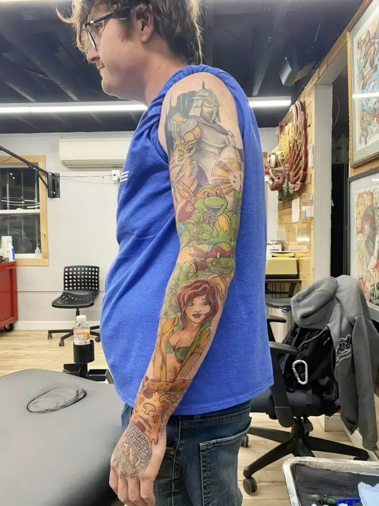 A person with glasses and a blue sleeveless shirt has a full sleeve tattoo featuring various colorful characters on their left arm.