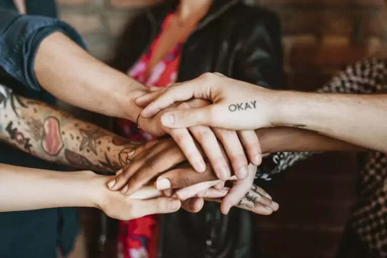 A group of people place their hands together in a stack, showing tattoos and different skin tones.