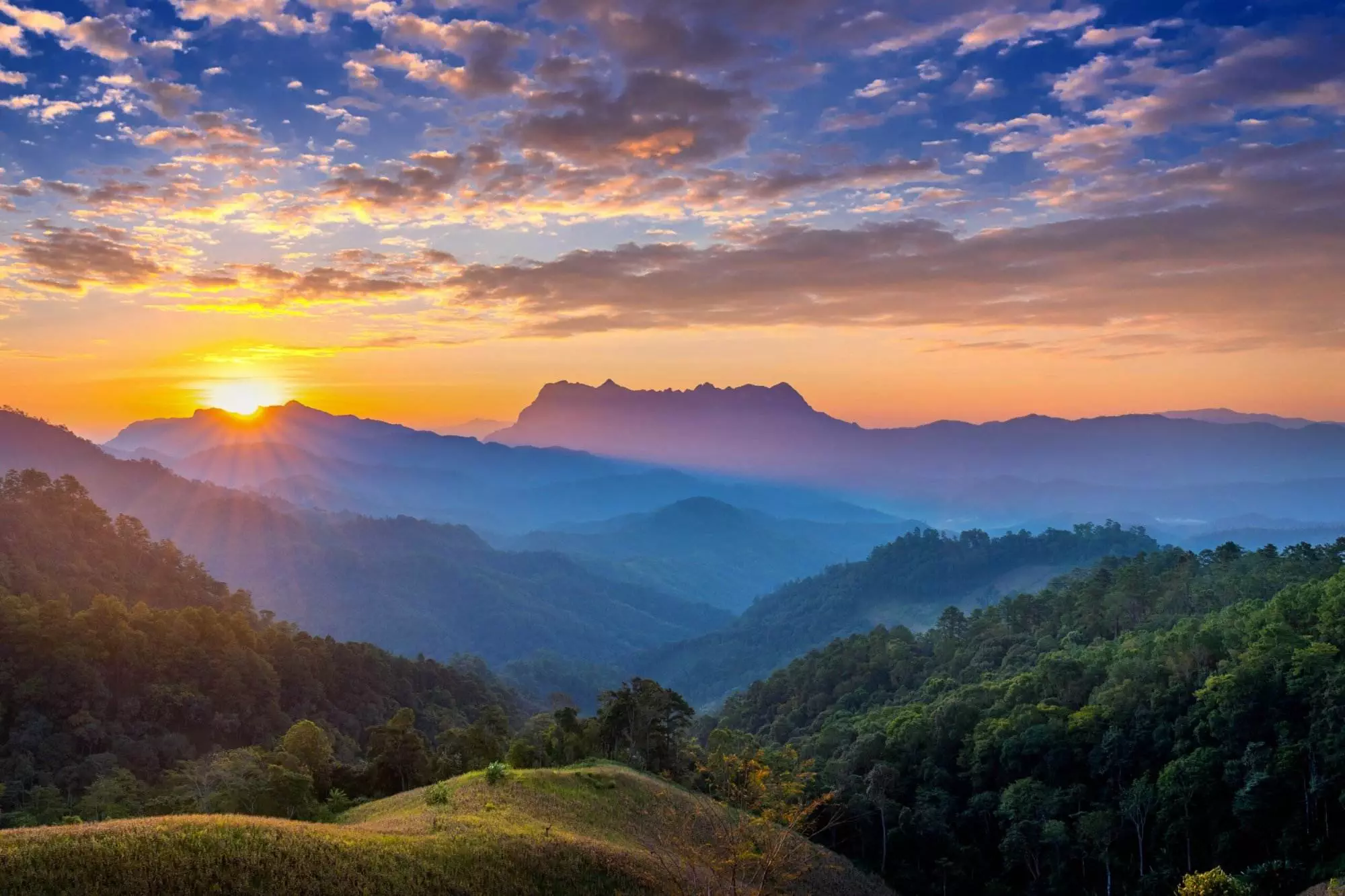 Sunrise over a mountain range with layers of blue-toned hills and a sky filled with clouds, casting a serene light on the valley below. Forested slopes are visible in the foreground.