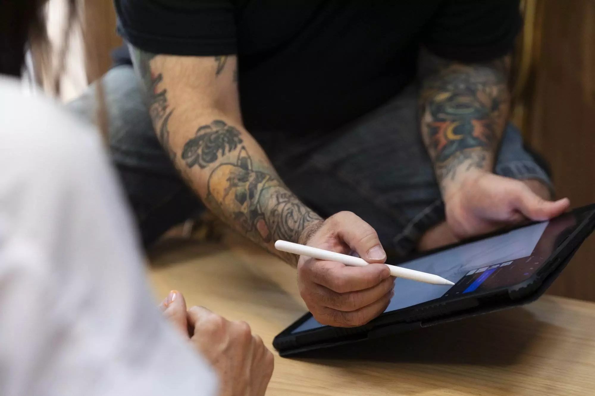 Person with tattoos on arms uses a stylus on a tablet, while another person watches from the foreground.