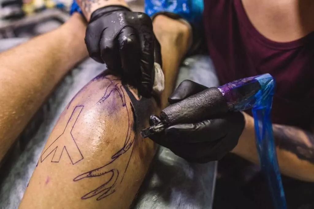 A tattoo artist with black gloves is using a tattoo machine to ink a design on a person's leg. The tattoo outline is partially completed.