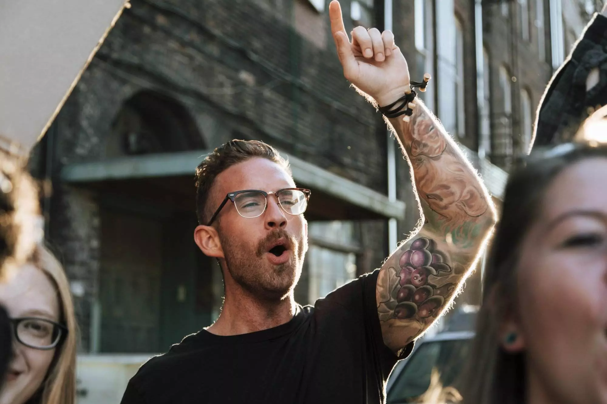 A man with glasses and tattoos is raising his arm, pointing upwards, and shouting. He is among a group of people.