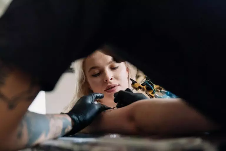 A tattoo artist wearing black gloves works on a tattoo on a woman's arm as she lies down and looks at the process.