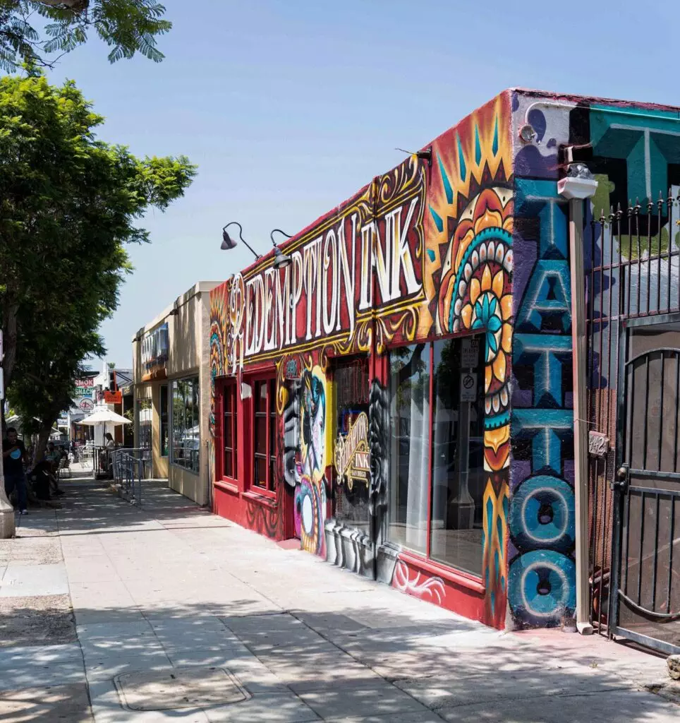 Street view of a colorful tattoo parlor with vibrant graffiti art on the storefront, surrounded by trees and neighboring shops.