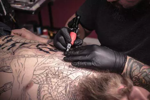 A tattoo artist in black gloves works on an intricate design on a person's chest.