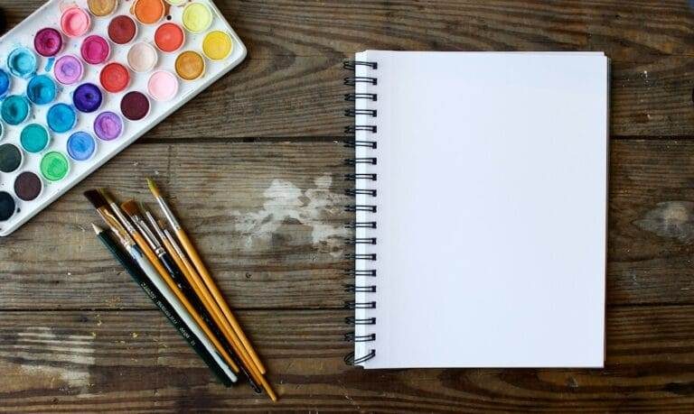 A spiral-bound blank sketchbook is placed on a wooden table next to a set of watercolor paints and various paintbrushes.