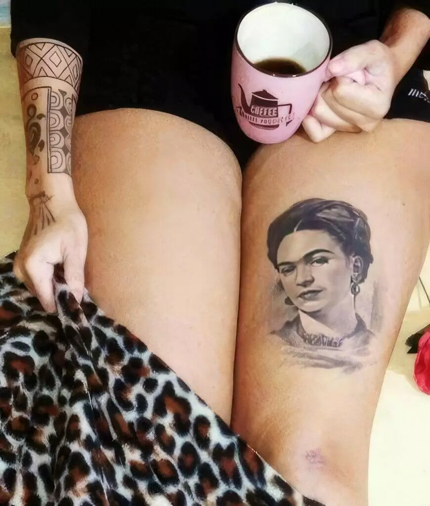 Person holding a pink mug with coffee while sitting with one leg covered by a leopard-print blanket, displaying a tattoo of a woman's face on the other thigh and a geometric arm tattoo.